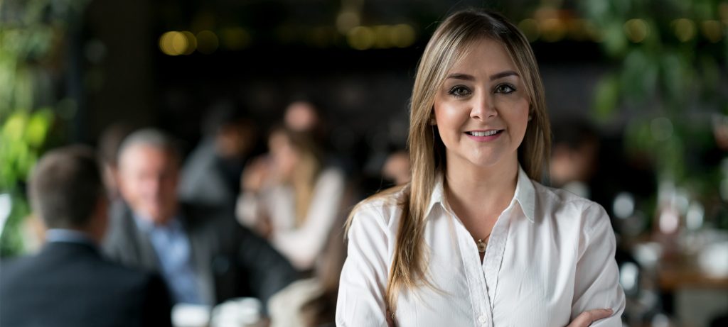 Photo d'une femme souriante debout à une terrasse où sont attablées des personnes